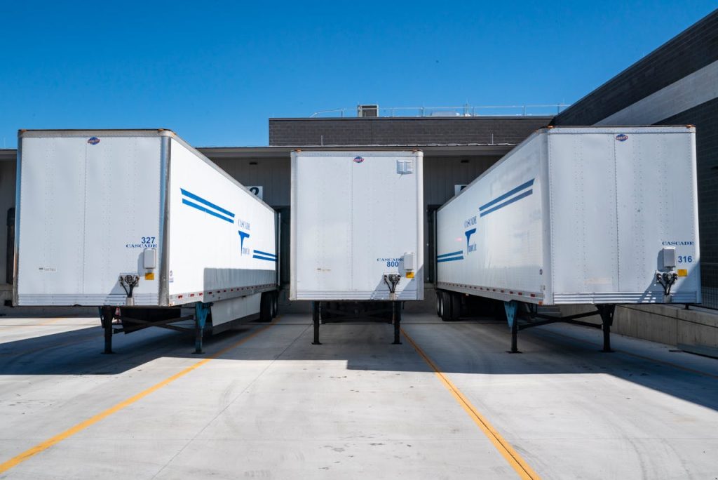 three-white-enclosed-trailers-1267325 Three white cargo trailers parked at an industrial shipping dock under clear blue skies.